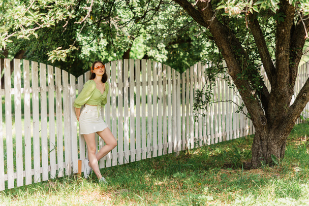 Young woman standing near fence and tree in summer park  - Φωτογραφία, εικόνα