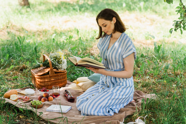 Πλευρική άποψη της γυναίκας ανάγνωση του βιβλίου κοντά στο φαγητό και το κρασί σε κουβέρτα στο πάρκο  - Φωτογραφία, εικόνα