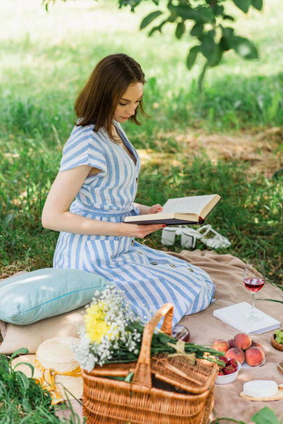 Mujer bonita leyendo libro cerca de vino, frutas y cesta en manta durante el picnic - Foto, imagen