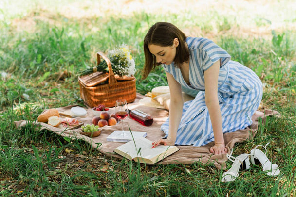 Woman in dress reading book near wine, fruits and blurred basket on blanket in park  - Φωτογραφία, εικόνα