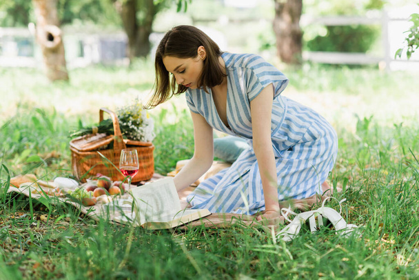 Vue latérale du livre de lecture de femme près du verre de vin pendant le pique-nique dans le parc  - Photo, image