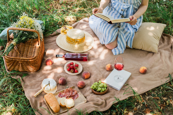 Vista recortada de la joven mujer sosteniendo libro durante el picnic con vino y comida en el parque  - Foto, Imagen