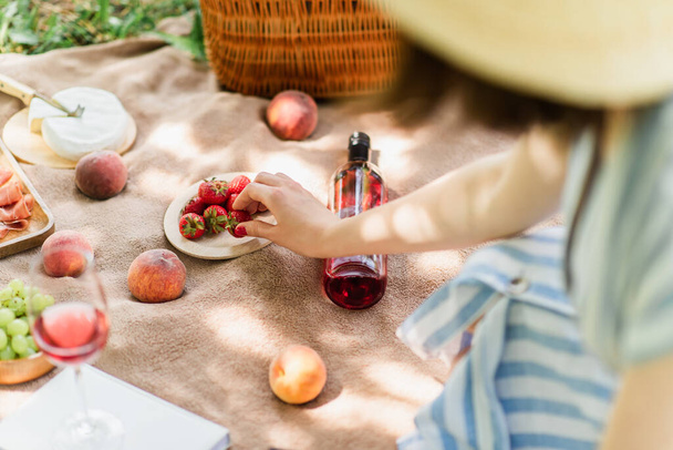 Vue recadrée de la femme prenant des fraises près du vin et des fruits sur couverture dans le parc  - Photo, image