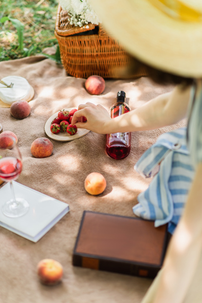 Bulanık kadın şarabın yanına çilek ve piknik battaniyesi üzerine kitap alıyor.  - Fotoğraf, Görsel