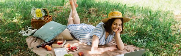 Щаслива жінка дивиться на ноутбук біля вина та їжі на ковдрі в парку, банер
  - Фото, зображення