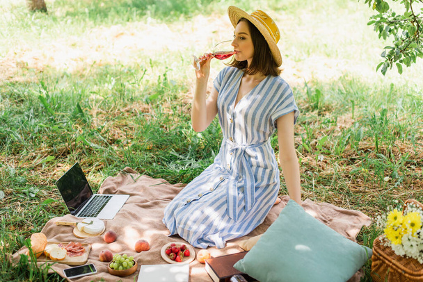 Pretty woman drinking wine near devices and food in park  - Φωτογραφία, εικόνα