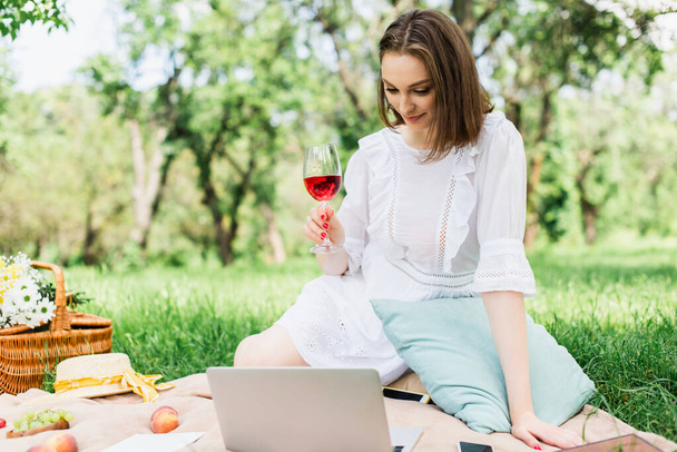 若い女性がピクニック毛布の上で機器や食べ物の近くにワインのガラスを保持  - 写真・画像