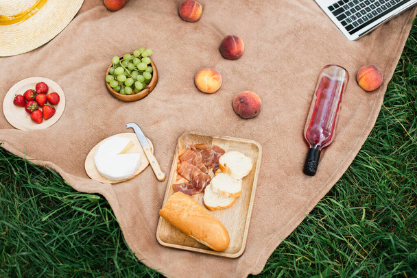 Vue de dessus de jambon savoureux, fruits avec vin et fromage près de l'ordinateur portable sur l'herbe  - Photo, image