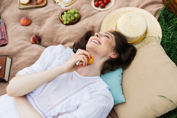 Top view of happy woman holding peach near pillows and sun hat on blanket  - Photo, Image