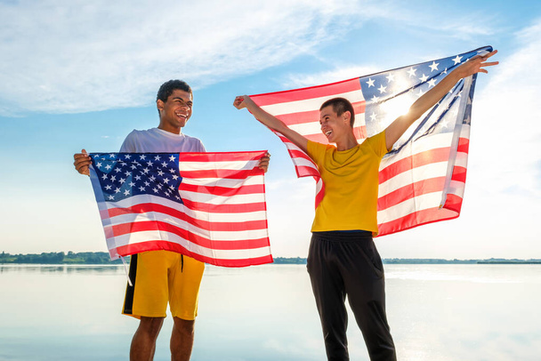 Δύο νεαροί χαρούμενοι και γελαστοί Αφρο-Αμερικανοί και Καυκάσιοι φίλοι, που υπερήφανα κρατούν αμερικανική σημαία ενάντια στον γαλάζιο ουρανό το ηλιοβασίλεμα το καλοκαίρι.  - Φωτογραφία, εικόνα