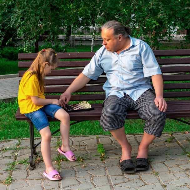 La niña juega al ajedrez con papá en el parque en un banco al aire libre. Día de ajedrez. Juegos educativos, educativos al aire libre. Desarrollo infantil. - Foto, imagen