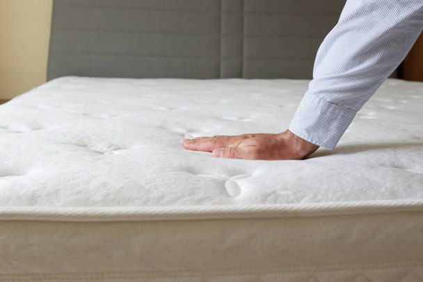 Cropped shot of man testing the firmness of white orthopedic mattress. Male pressing hypoallergenic foam mattresses surface to check its softness. Close up, copy space, top view, background. - Photo, Image