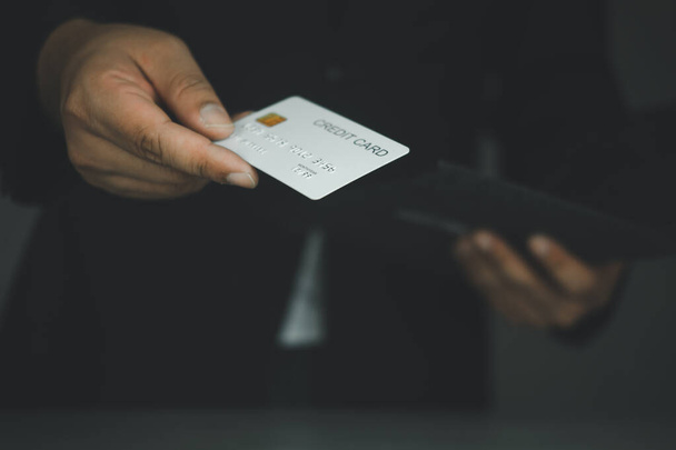 Empresário em um terno clássico preto segurando um cartão de crédito de plástico branco no estúdio em um fundo escuro. conceito de compras - Foto, Imagem
