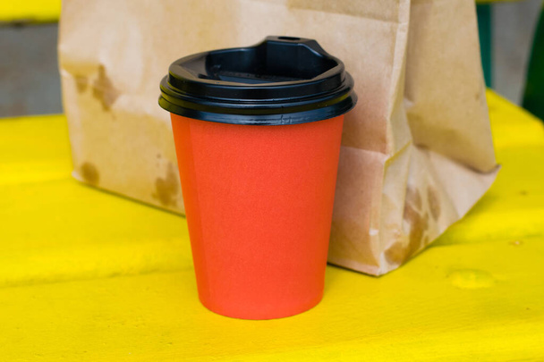 Piros papír csésze kávé és papírzacskó étellel egy sárga fa padon, a strandon vagy a parkban egy nyári napsütéses napon. - Fotó, kép