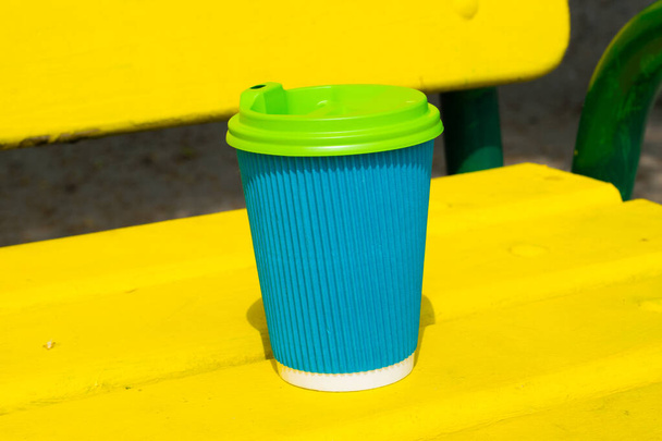 Tazza di carta di colore blu con un caffè per andare su una panchina di legno giallo sulla spiaggia o nel parco nella giornata di sole estiva - Foto, immagini