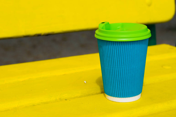 Papieren beker blauwe kleur met een koffie te gaan op gele houten bank op het strand of in het park in de zomer zonnige dag - Foto, afbeelding