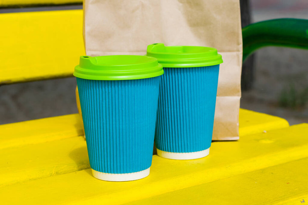 Blaue Pappbecher für Kaffee und eine Papiertüte mit Lebensmitteln auf einer gelben Holzbank, am Strand oder im Park an einem sonnigen Sommertag - Foto, Bild
