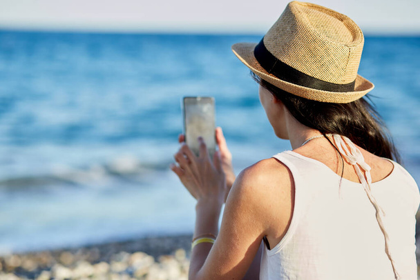 Young brunette with a phone on a sunbed under a beach umbrella on the beach near the sea. Vacation concept, taking a selfie, chatting with friends or freelancer, working remotely, webinar - Photo, Image