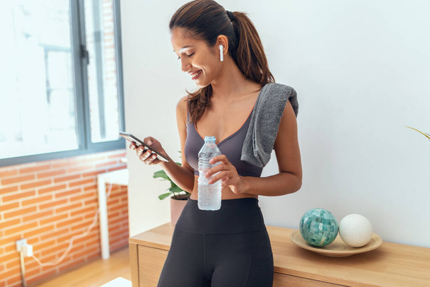 Shot of sporty young woman sending messages with her smart phone while take a break of exercise standing in living room at home. - Foto, Imagem