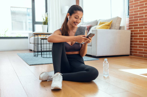 Shot of sporty young woman using her mobile phone while holding water bottle after a pilates class on the floor at home. - Valokuva, kuva