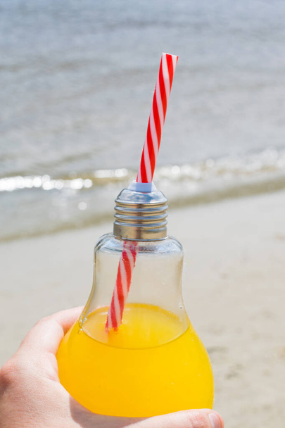 Frisch oder Cocktail in einer Glasflasche mit einem Strohhalm in weiblicher Hand vor dem Hintergrund des Strandes und des Wassers, vertikales Format. Reise- und Freizeitkonzept - Foto, Bild