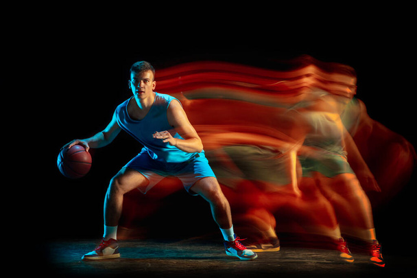 Professional male basketball player in blue uniform playing basketball isolated over dark studio background in mixed light. Concept of healthy lifestyle, professional sport, hobby. - Fotografie, Obrázek