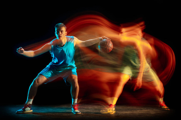 Young caucasian male basketball player playing basketball isolated over dark studio background in mixed light. Concept of healthy lifestyle, professional sport, hobby. - Valokuva, kuva