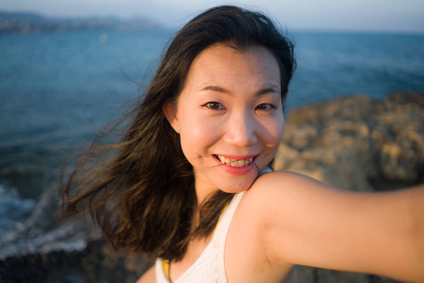 Genç ve güzel Asyalı kadın deniz kenarında cep telefonuyla selfie çekiyor. Koreli kız yaz tatilinin tadını çıkarıyor. Plajda rahatça fotoğraf çekiyor. - Fotoğraf, Görsel