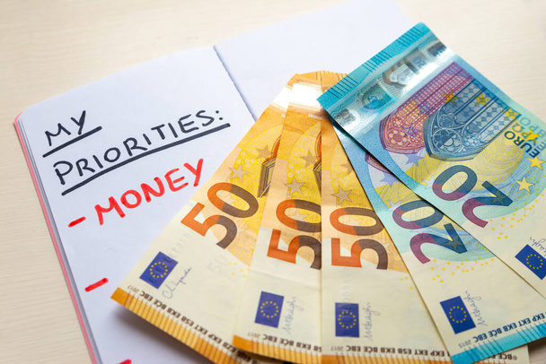 Κενή σελίδα ημερολογίου, με κείμενο "Προτεραιότητες" με μαύρο μελάνι και κείμενο "Χρήματα" με κόκκινο μελάνι, με τραπεζογραμμάτια ευρώ δίπλα του. Πλούτος και σκοπός στη ζωή.  - Φωτογραφία, εικόνα