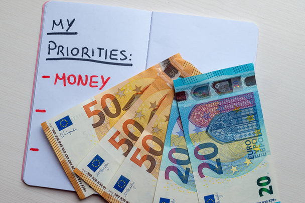 Página de diario en blanco, con texto "Prioridades" en tinta negra; y texto "Dinero" en tinta roja, con billetes en euros a su lado. Riqueza y propósito en la vida.  - Foto, imagen
