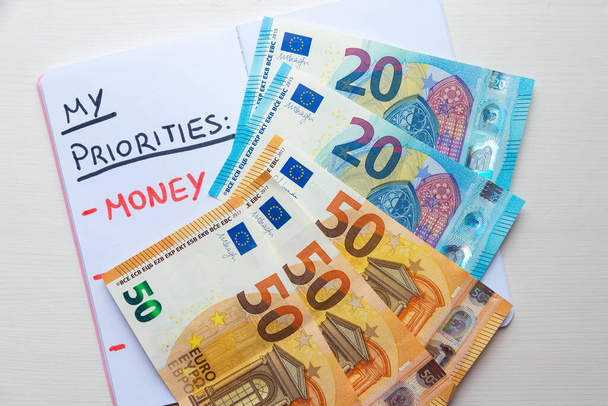 Κενή σελίδα ημερολογίου, με κείμενο "Προτεραιότητες" με μαύρο μελάνι και κείμενο "Χρήματα" με κόκκινο μελάνι, με τραπεζογραμμάτια ευρώ δίπλα του. Πλούτος και σκοπός στη ζωή.  - Φωτογραφία, εικόνα