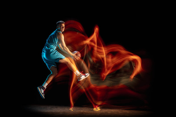 Young caucasian male basketball player playing basketball isolated over dark studio background in mixed light. Concept of healthy lifestyle, professional sport, hobby. - Фото, зображення