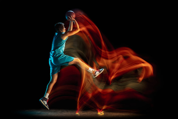 Young caucasian male basketball player playing basketball isolated over dark studio background in mixed light. Concept of healthy lifestyle, professional sport, hobby. - Fotó, kép