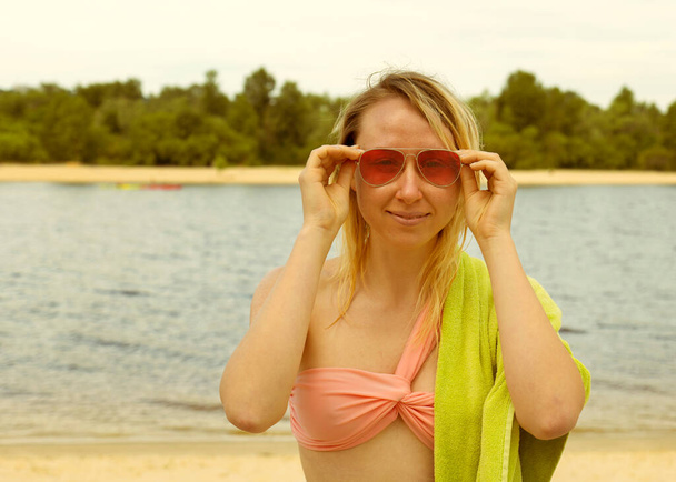Portrét mladé blondýny v červených brýlích s ručníkem u řeky. Dívka odpočívající na pláži - Fotografie, Obrázek