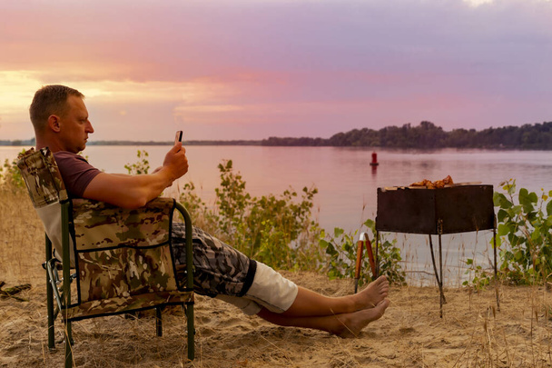 Man met mobiele telefoon in de hand is genieten van een prachtig landschap, terwijl zitten op het strand en grillen een barbecue - Foto, afbeelding