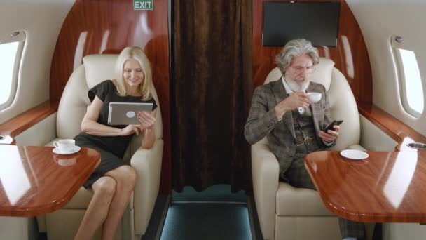 Casal rico maduro usando smartphone e tablet pc enquanto luxo viajando em avião a jato privado. Homem sênior e mulher usando gadgets, olhando para a janela enquanto voa em companhias aéreas de primeira classe - Filmagem, Vídeo