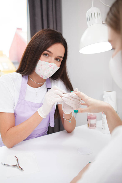 Vertical portrait of a professional manicurist wearing face mask looking to the camera while working - Foto, Imagem