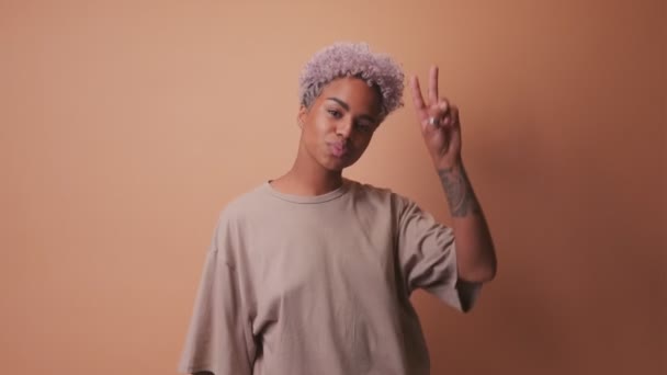 Friendly African American woman makes peace sign over eye over beige background. - Кадры, видео