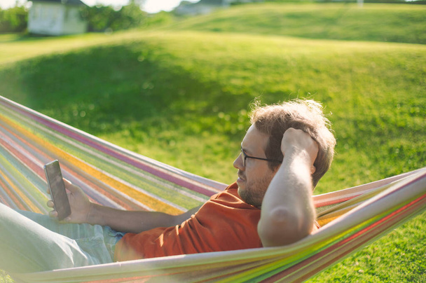 portrait of attractive nerd   man with glasses in the park with green lawn have a nice sunset in  the hammock  . Happy procrastination with phone - Fotó, kép
