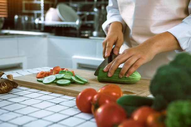 Femme utilisant le couteau et les mains coupant le concombre sur le panneau en bois dans la salle de cuisine. - Photo, image