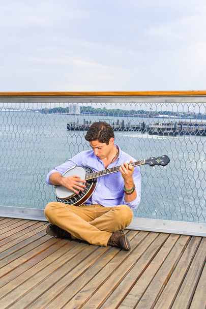 Опускаючи голову, перетинаючи ноги і думаючи, молодий музикант сидить на палубі, граючи банджо
 - Фото, зображення
