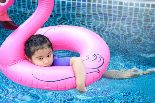 Αξιολάτρευτο κορίτσι από την Ασία που κολυμπάει με ροζ φλαμίνγκο πλαστικό παιχνίδι σε μπλε νερά, χαριτωμένο παιδί που παίζει σε φουσκωτούς σωλήνες σε μια πισίνα σε μια ηλιόλουστη μέρα, διασκεδάζοντας στις καλοκαιρινές διακοπές.  - Φωτογραφία, εικόνα
