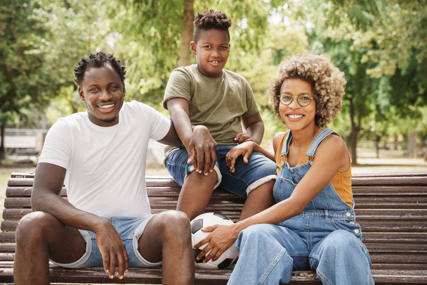 Portrait of happy African American family with young cool millennial parents and cute smiling kid sitting on bench in outdoor public park - Фото, изображение