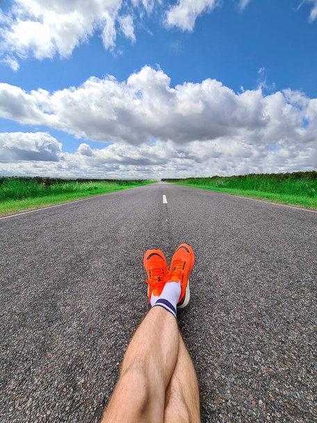 青い空を背景に道路上に横たわるスポーツスニーカーの男性の足と雲のクローズアップ - 写真・画像