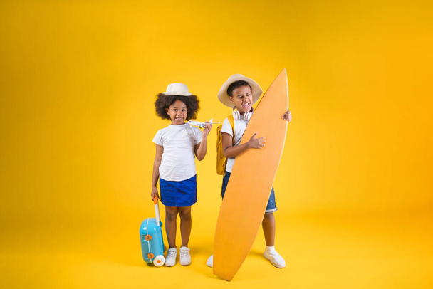 Happy sister and brother standing near surfboard with backpack and suitcase on yellow background - Фото, изображение