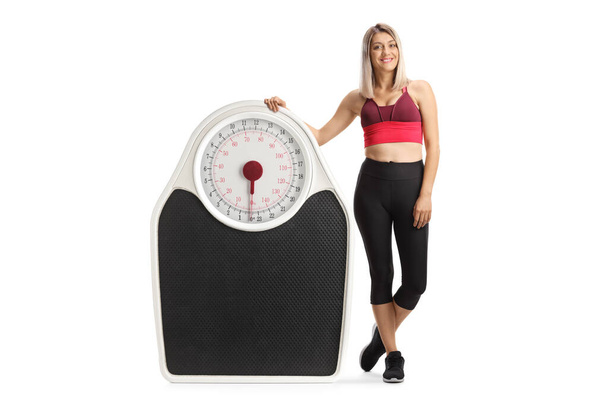 Full length portrait of a young woman in sports clothing leaning on a big weight scale isolated on white background - Zdjęcie, obraz
