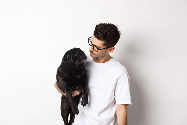 Handsome young man holding his black pug, dot and pet owner looking each other faces, standing against white background - Photo, Image