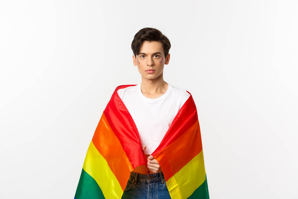 Image of serious-looking gender fluid person with glitter on face, wearing lgbtq rainbow flag, standing over white background - Photo, Image