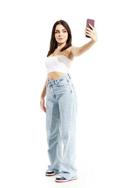 Zeki telefonuyla selfie çeken oldukça kendine güvenen, trend bir Z genci genç kadın. Beyaz arkaplanda tam vücut portresi izole edildi - Fotoğraf, Görsel