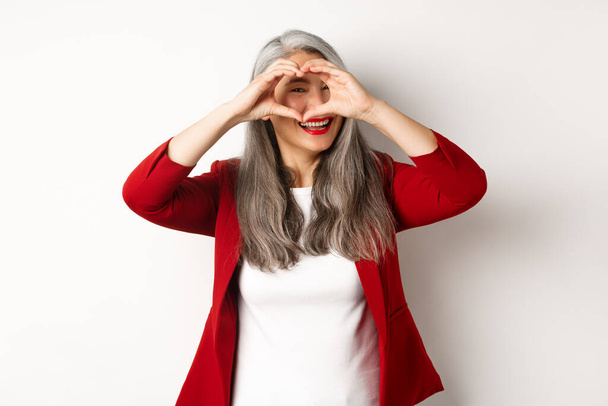 Lovely asian middle-aged woman with grey hair, wearing red blazer, showing heart sign and peeking throught it, I love you gesture, standing over white background - Photo, Image
